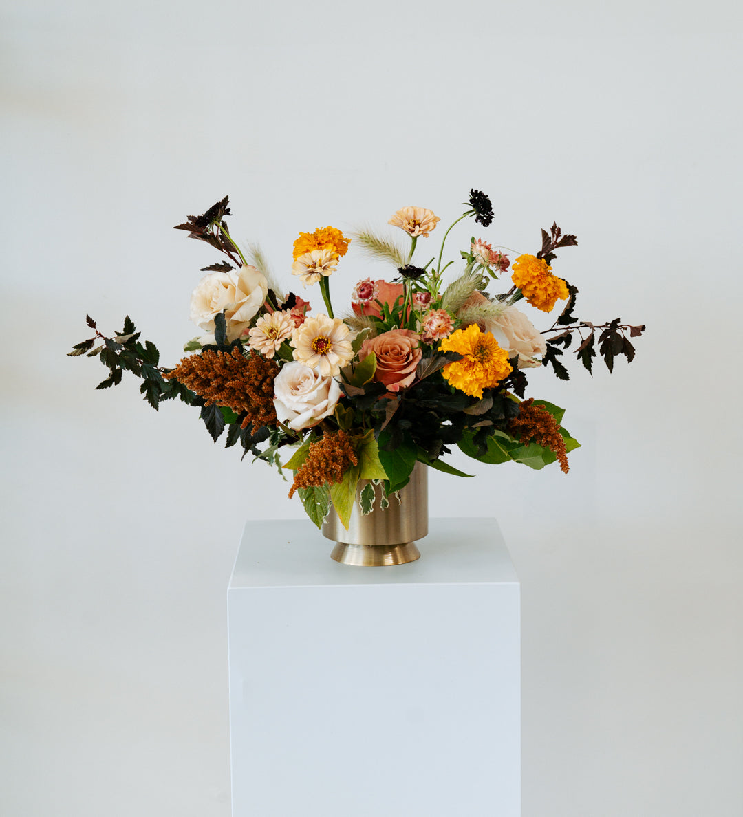 Lush, fall themed Floral arrangement in a gold vase on a white pedestal against a white background