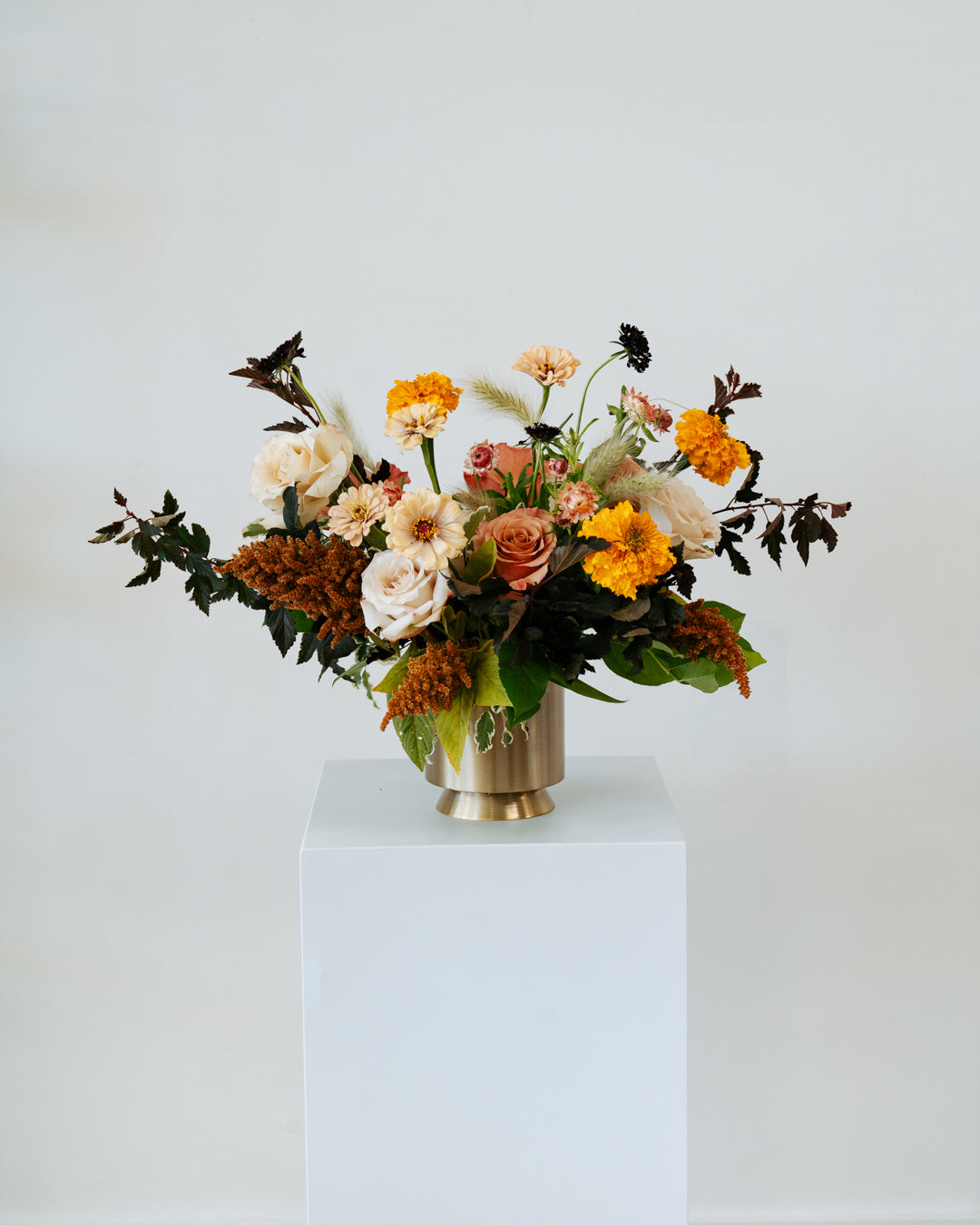 Lush, fall themed Floral arrangement in a gold vase on a white pedestal against a white background
