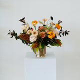 Lush, fall themed Floral arrangement in a gold vase on a white pedestal against a white background