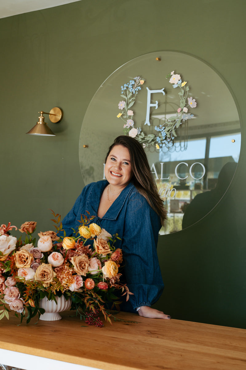 Rachel Priest stands smiling in front of a colorful vase filled with fresh flowers.