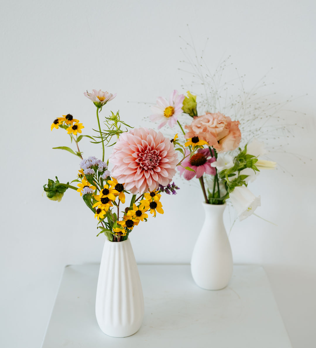 Two white ceramic vases filled with a mixed floral arrangement on a white pedestal against a white wall made by Flocal Co in Columbus Ohio