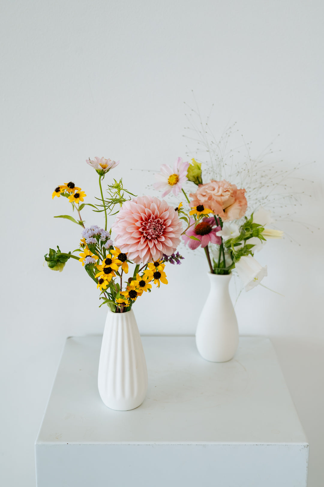 Two white ceramic vases filled with a mixed floral arrangement on a white pedestal against a white wall made by Flocal Co in Columbus Ohio
