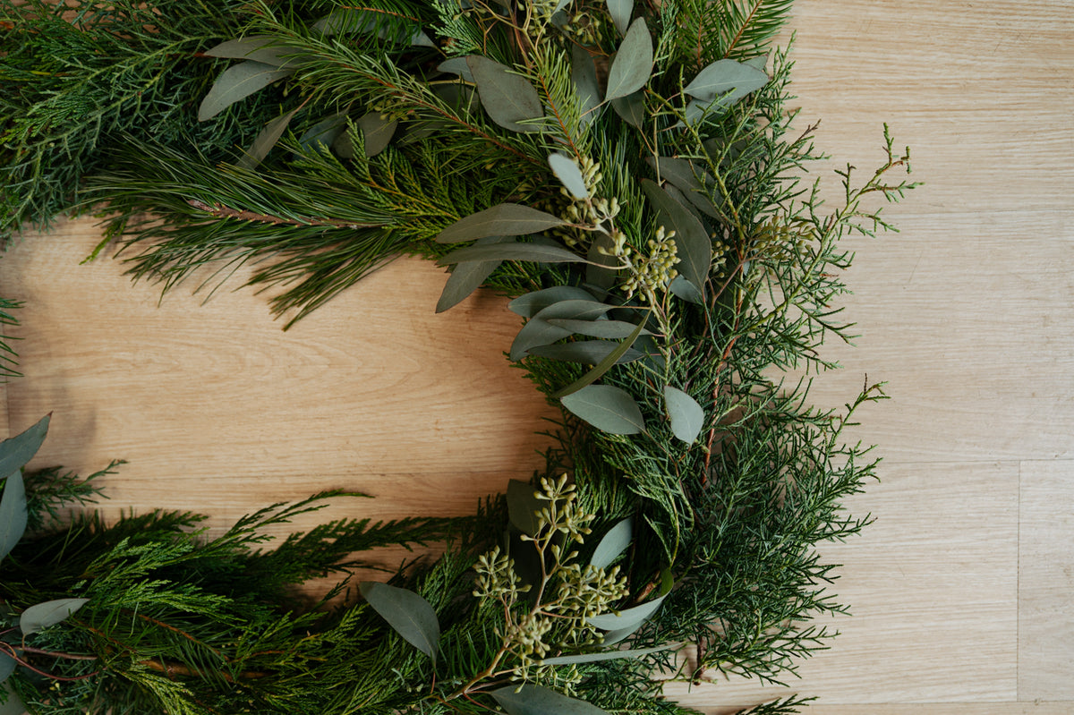 A lush evergreen wreath with eucalyptus leaves and tiny berries frames a blank oval wooden plaque on a pale wood background, creating a calm, rustic feel.
