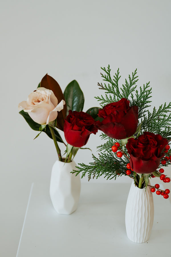 Two white ceramic vases with floral arrangements on a white surface against a light gray background. The arrangement on the left features a pale blush rose and green leaves, while the arrangement on the right features three deep red roses with green pine branches and small red berries.