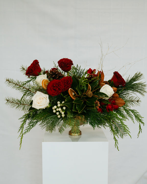 A lush floral centerpiece of red and white roses, greenery, berries, and dried branches sits on a white pedestal, creating an elegant, festive mood.