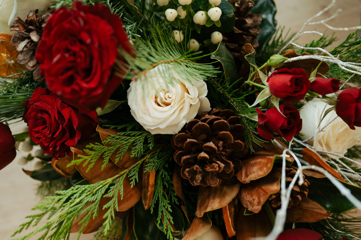 A lush winter bouquet of red and ivory roses, pinecones, evergreen sprigs, white berries, and dried leaves conveys a warm, festive, rustic mood.