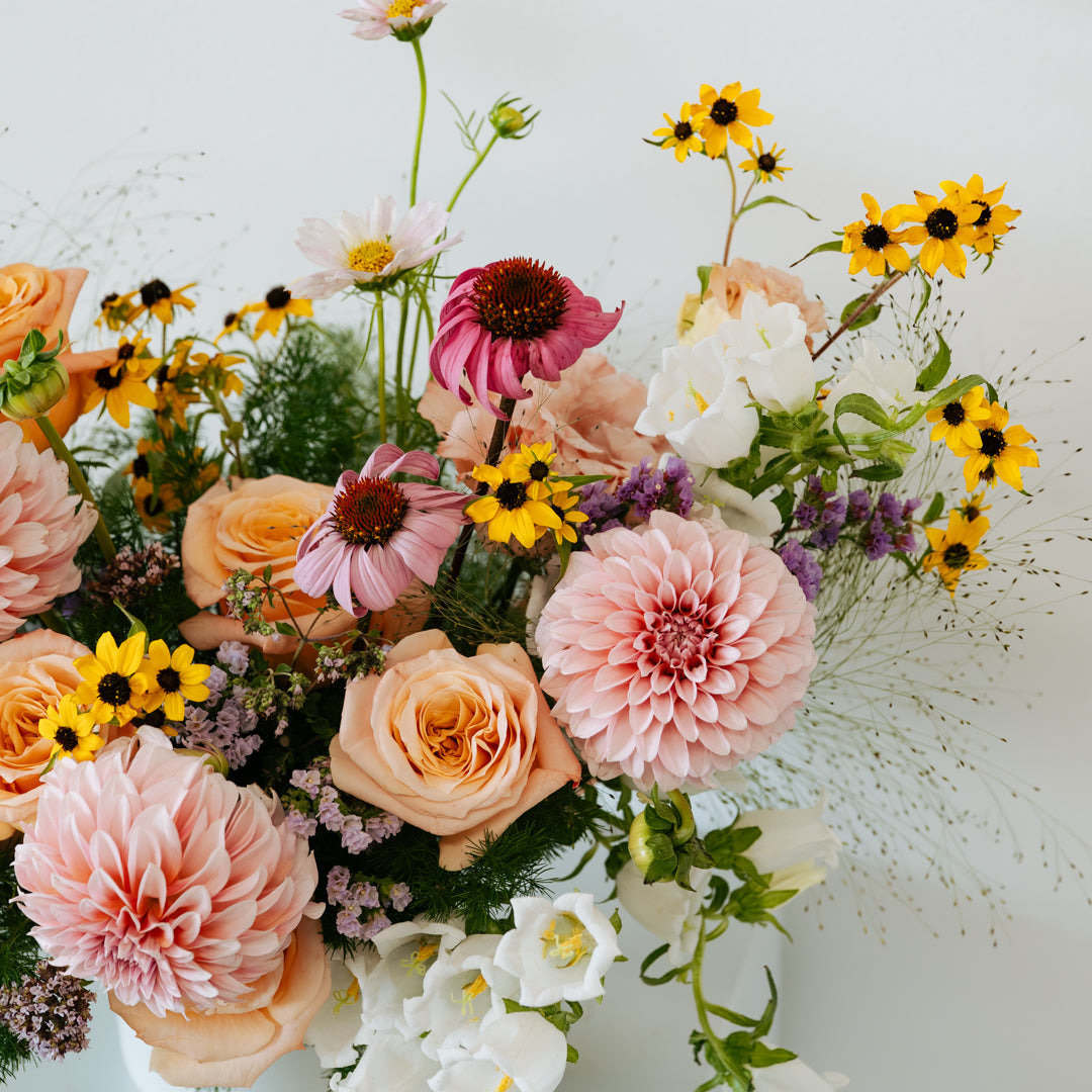 A vibrant mixed floral arrangement in a white vase, featuring large pink dahlias, peach roses, yellow sunflowers, purple coneflowers, and white blossoms against a clean white background made by Flocal Co in Columbus, Ohio