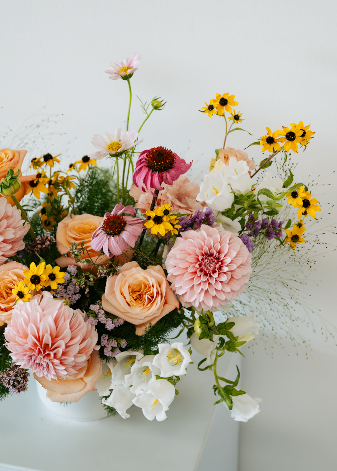 A vibrant mixed floral arrangement in a white vase, featuring large pink dahlias, peach roses, yellow sunflowers, purple coneflowers, and white blossoms against a clean white background made by Flocal Co in Columbus, Ohio
