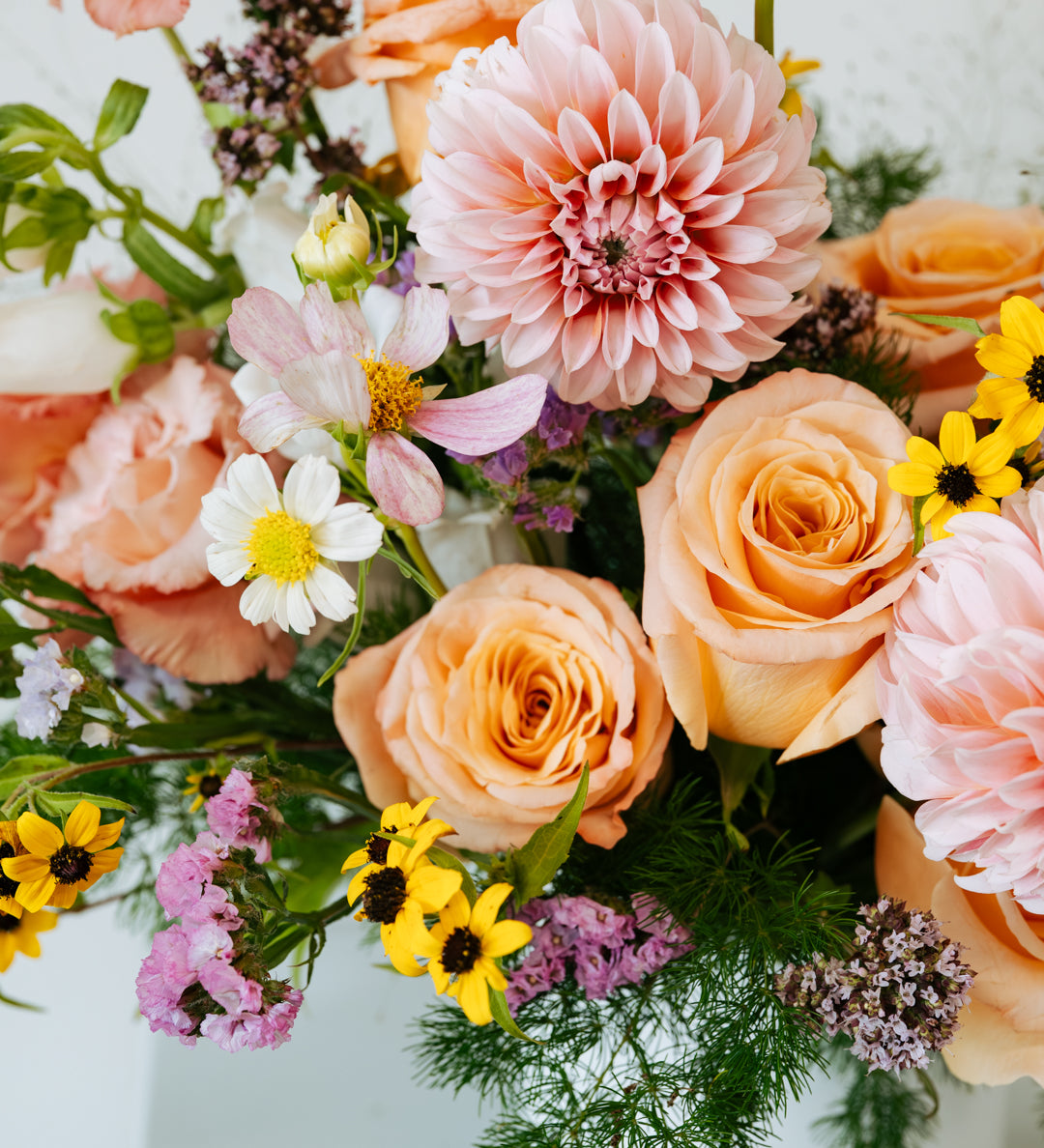 A close-up of a pastel flower arrangement featuring peach and pink roses, a large pink dahlia, white cosmos, yellow floral accents and green foliage in a white vase made by Flocal Co in Columbus, Ohio