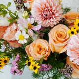 A close-up of a pastel flower arrangement featuring peach and pink roses, a large pink dahlia, white cosmos, yellow floral accents and green foliage in a white vase made by Flocal Co in Columbus, Ohio