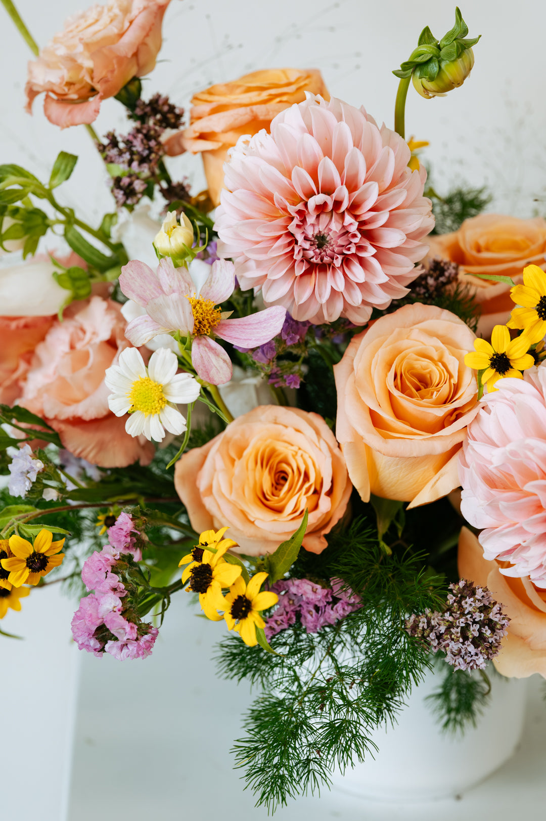 A close-up of a pastel flower arrangement featuring peach and pink roses, a large pink dahlia, white cosmos, yellow floral accents and green foliage in a white vase made by Flocal Co in Columbus, Ohio