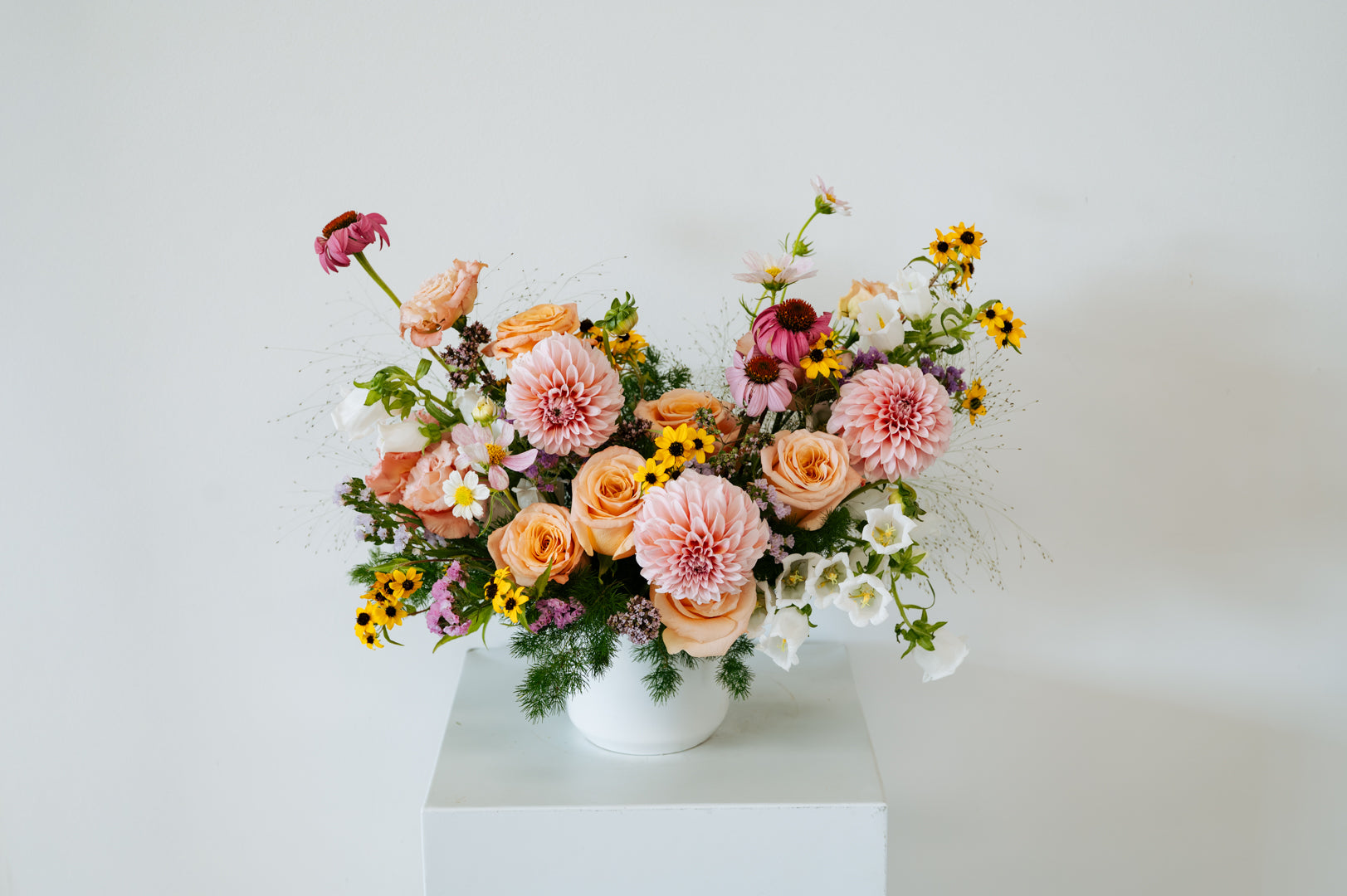 A vibrant floral arrangement featuring pink, peach, yellow, and white flowers in a white ceramic pot, displayed on a white pedestal against a clean white background made by Flocal Co in Columbus Ohio