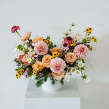 A vibrant floral arrangement featuring pink, peach, yellow, and white flowers in a white ceramic pot, displayed on a white pedestal against a clean white background made by Flocal Co in Columbus Ohio