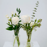 Two clear glass vases with white flowers on a white surface and light gray background at Flocal Co.