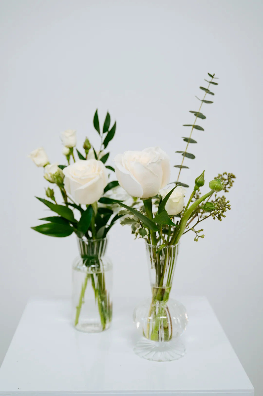Two clear glass vases with white flowers on a white surface and light gray background at Flocal Co.