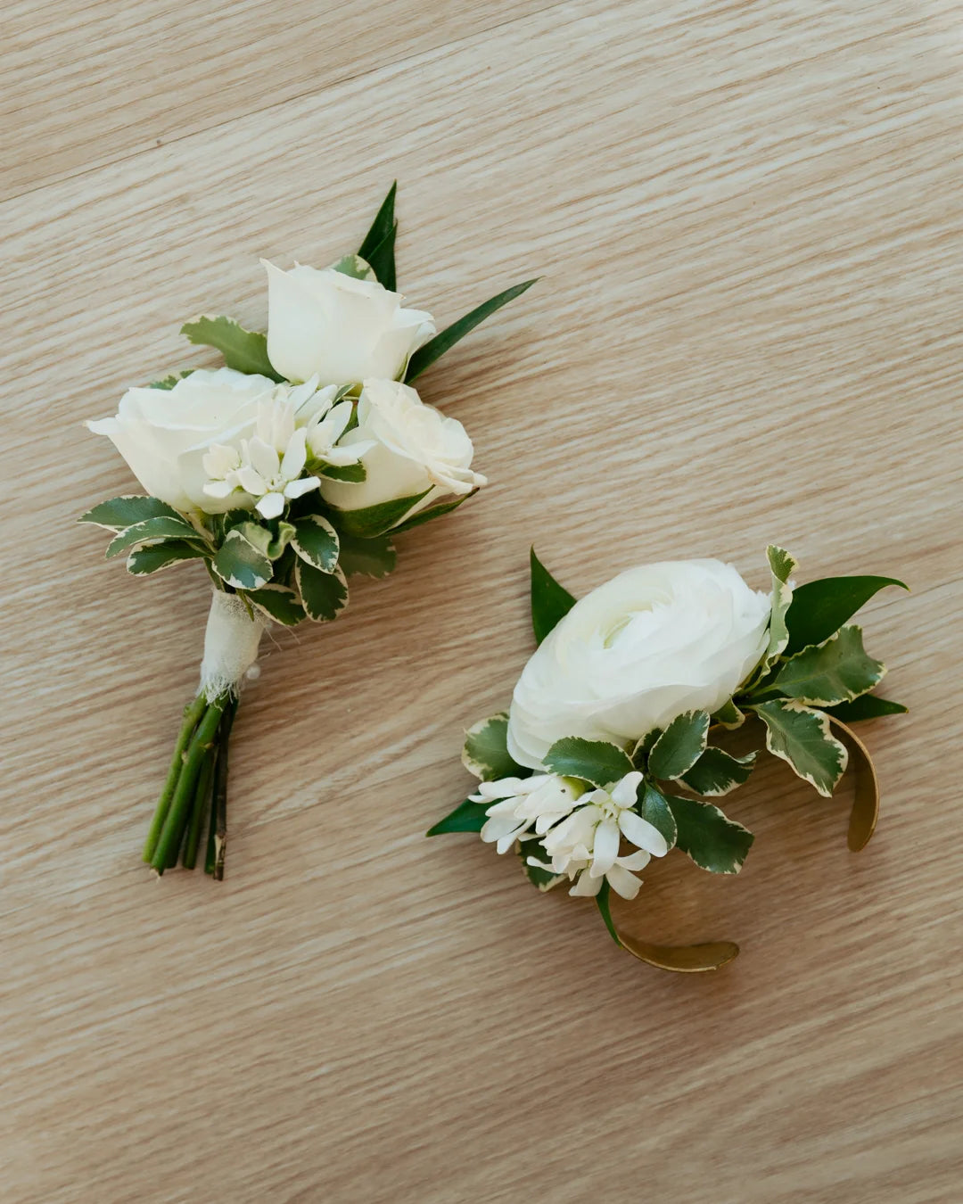 Two white floral corsages by Flocal Co. with green leaves on a wooden surface