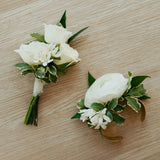Two white floral corsages by Flocal Co. with green leaves on a wooden surface