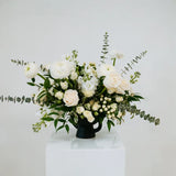 White and green floral arrangement on a white pedestal against a light gray background