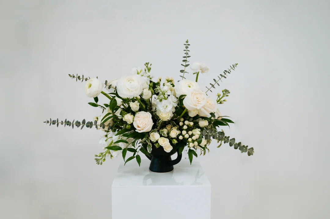 White and green floral arrangement on a white pedestal against a light gray background