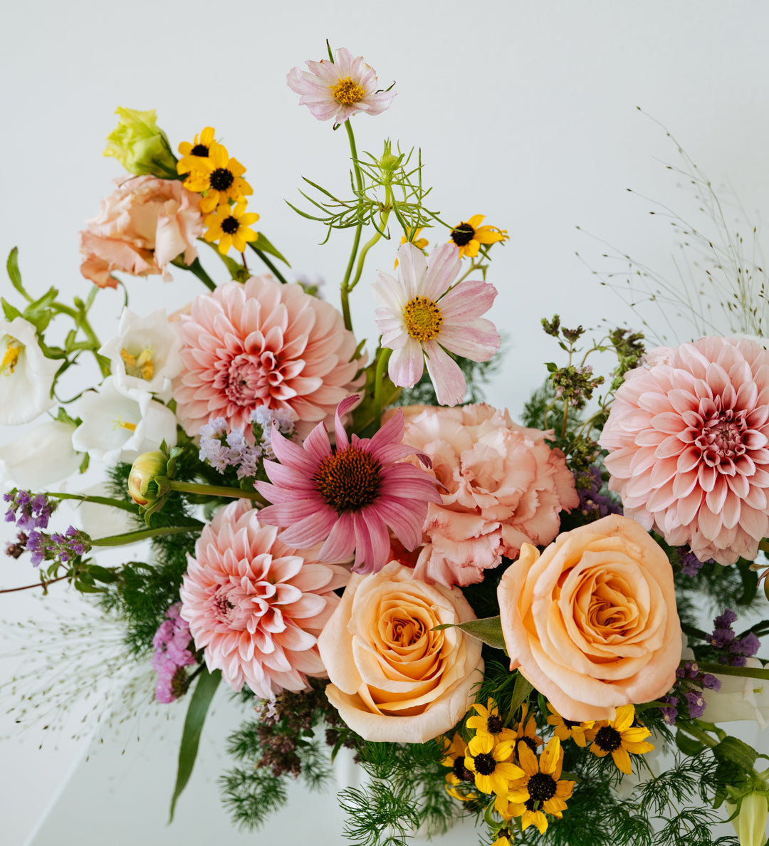 A vibrant mixed floral arrangement featuring peach roses, pink dahlias, yellow sunflowers, and green foliage, presented on a clean white surface against a white background made by Flocal Co in Columbus, Ohio