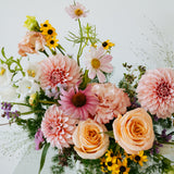 A vibrant mixed floral arrangement featuring peach roses, pink dahlias, yellow sunflowers, and green foliage, presented on a clean white surface against a white background made by Flocal Co in Columbus, Ohio
