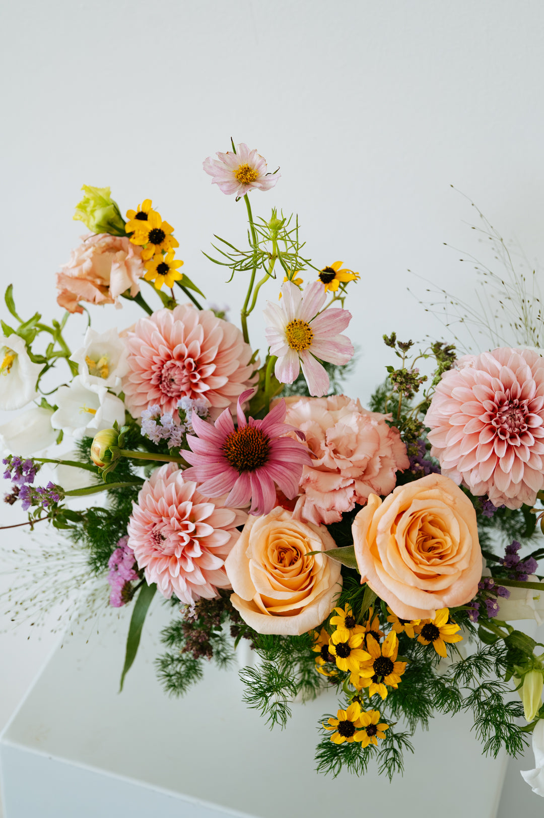 A vibrant mixed floral arrangement featuring peach roses, pink dahlias, yellow sunflowers, and green foliage, presented on a clean white surface against a white background made by Flocal Co in Columbus, Ohio
