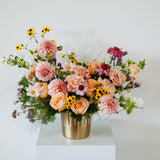 A vibrant floral arrangement featuring peach roses, pink chrysanthemums, yellow daisies, and purple coneflowers in a metallic gold bucket vase, displayed on a white pedestal against a light gray background made by Flocal Co in Columbus Ohio