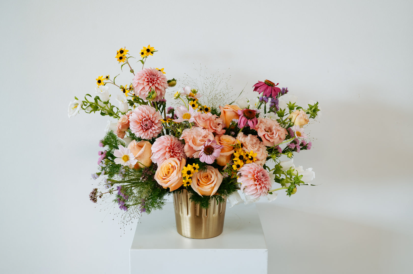 A vibrant floral arrangement featuring peach roses, pink chrysanthemums, yellow daisies, and purple coneflowers in a metallic gold bucket vase, displayed on a white pedestal against a light gray background made by Flocal Co in Columbus Ohio