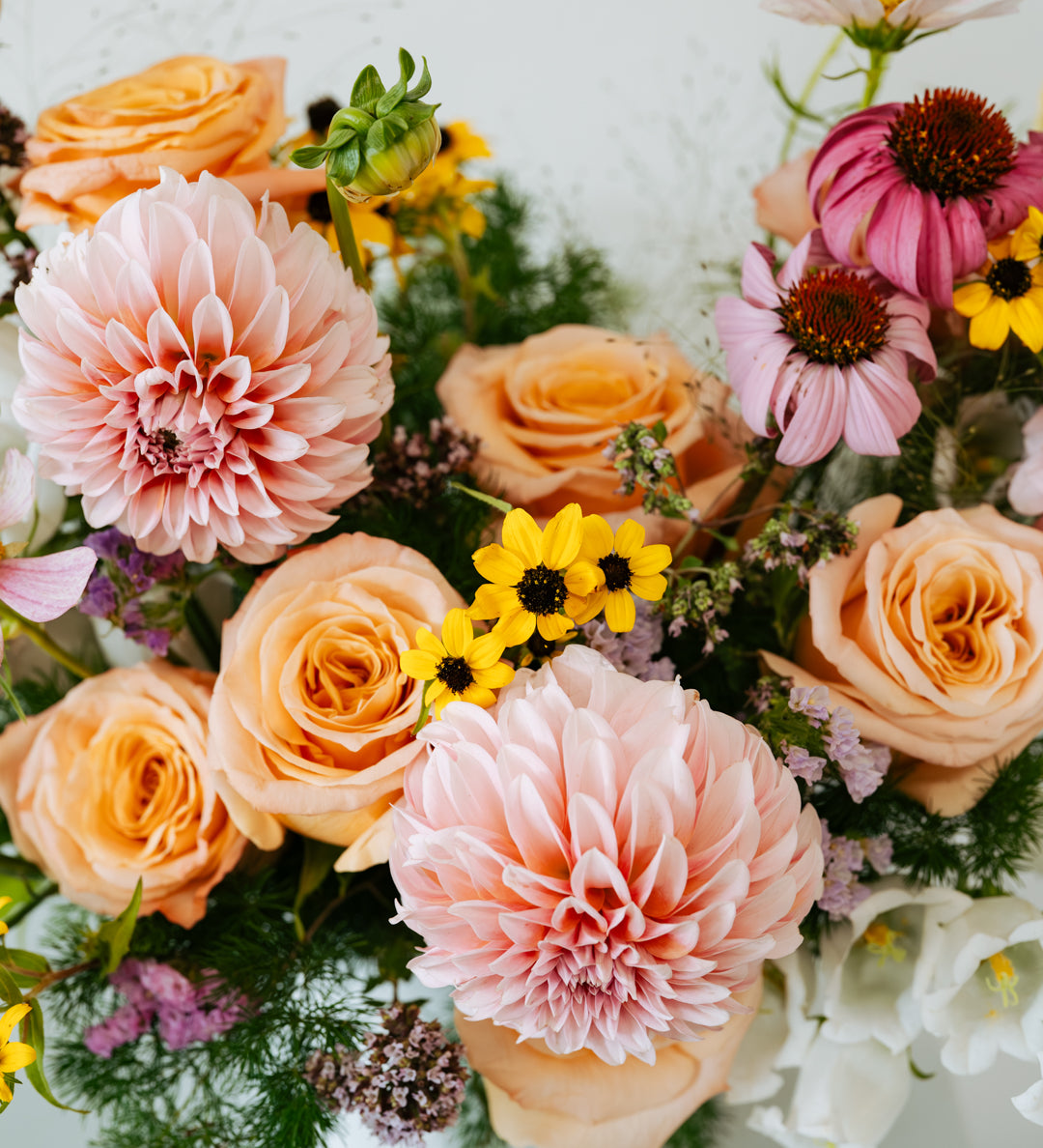 A close-up floral arrangement featuring peach and pink roses, large pale pink dahlias, pink coneflowers, small yellow daisy-like flowers, and green foliage on a light neutral background made by Flocal Co in Columbus Ohio 