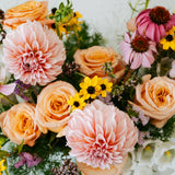 A close-up floral arrangement featuring peach and pink roses, large pale pink dahlias, pink coneflowers, small yellow daisy-like flowers, and green foliage on a light neutral background made by Flocal Co in Columbus Ohio 