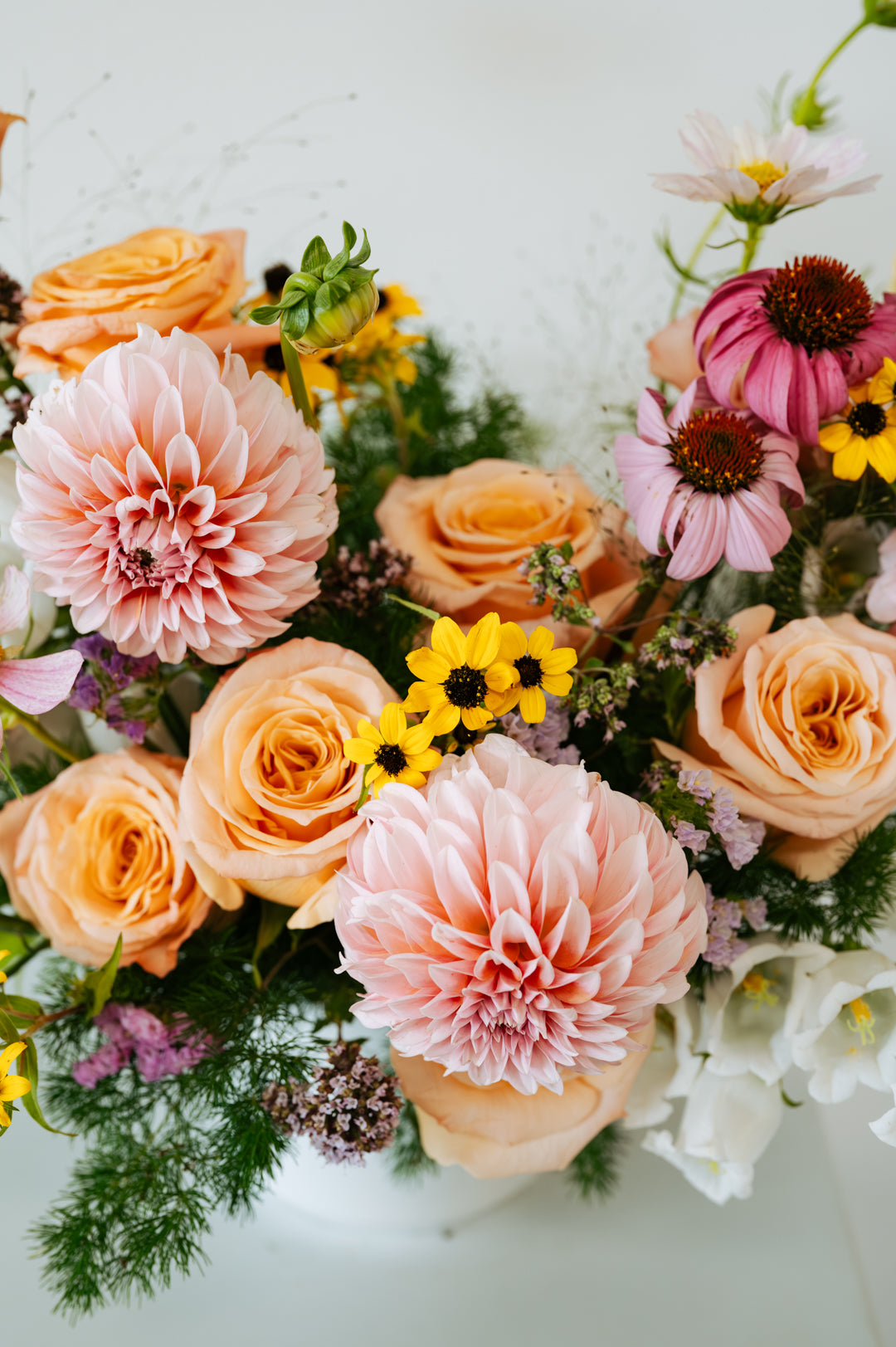 A close-up floral arrangement featuring peach and pink roses, large pale pink dahlias, pink coneflowers, small yellow daisy-like flowers, and green foliage on a light neutral background made by Flocal Co in Columbus Ohio 
