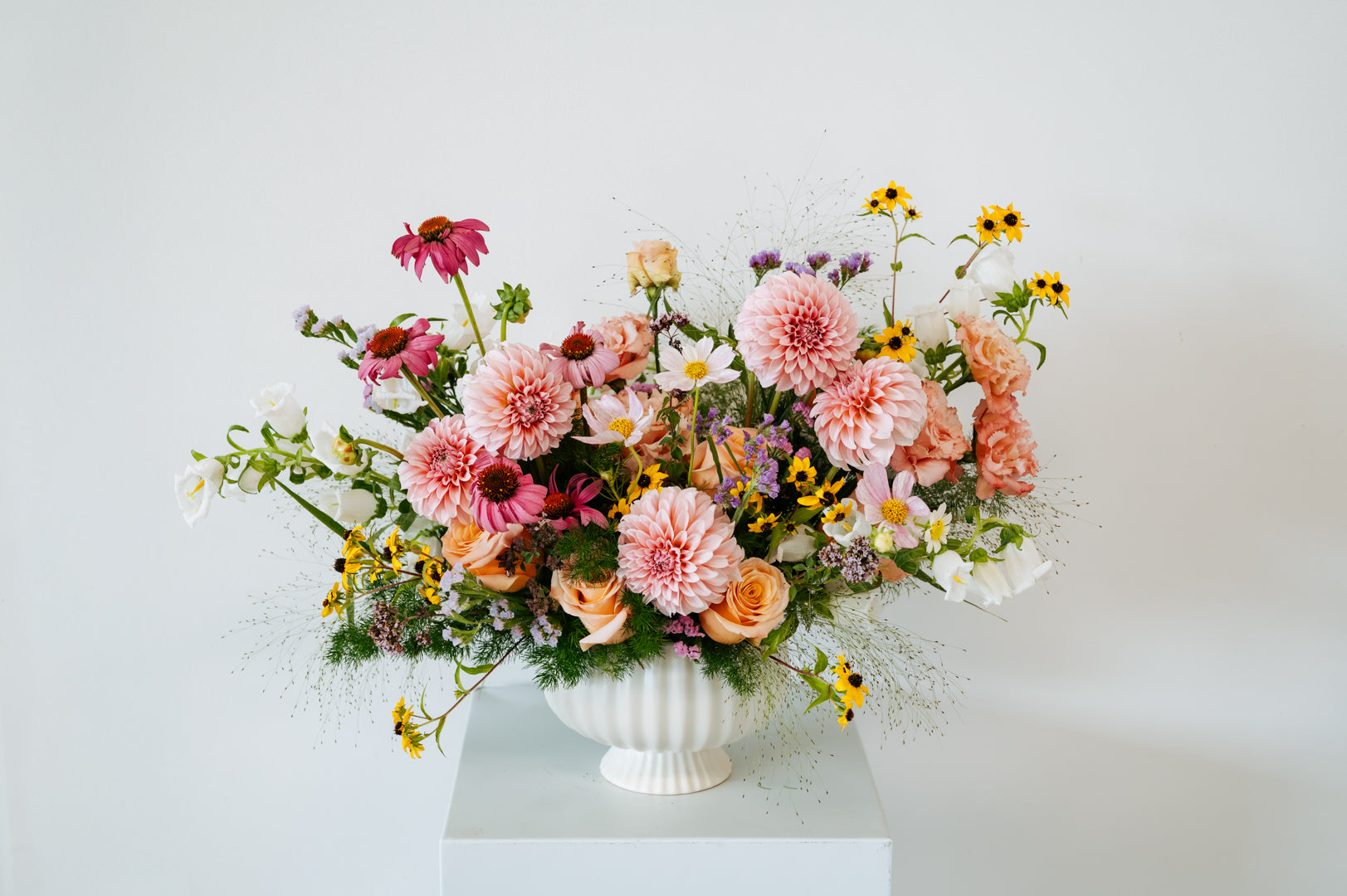 A vibrant wildflower bouquet in a white ribbed ceramic urn, displayed on a white pedestal against a clean white background made by Flocal Co in Columbus, Ohio