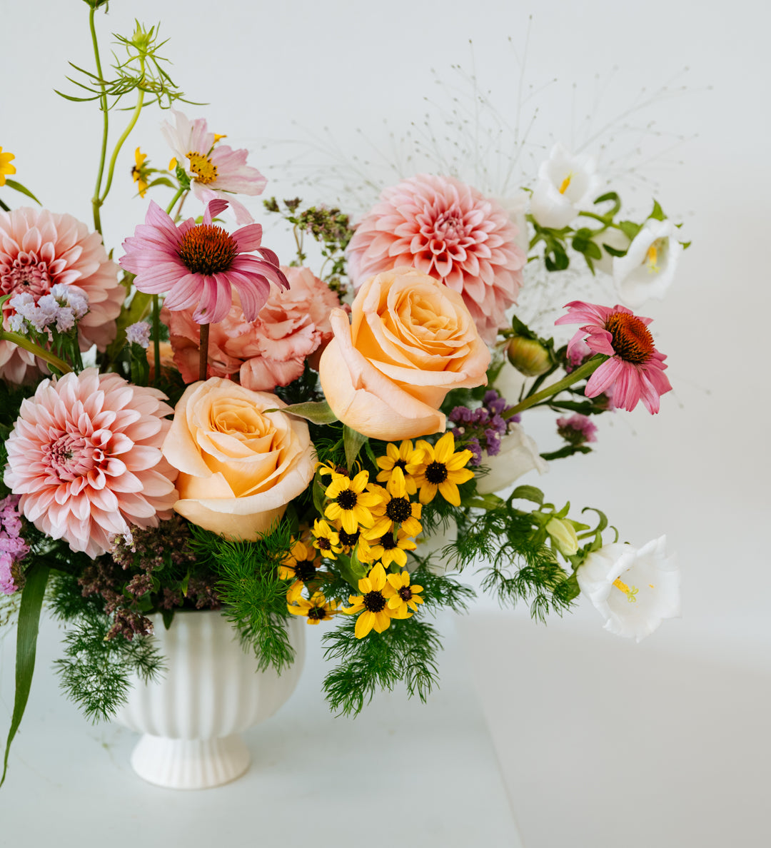 A pastel floral arrangement in a white ribbed ceramic vase, featuring peach roses, pink dahlias, yellow daisies, and green foliage, displayed on a white pedestal against a white background made by Flocal Co in Columbus, Ohio