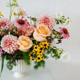 A pastel floral arrangement in a white ribbed ceramic vase, featuring peach roses, pink dahlias, yellow daisies, and green foliage, displayed on a white pedestal against a white background made by Flocal Co in Columbus, Ohio
