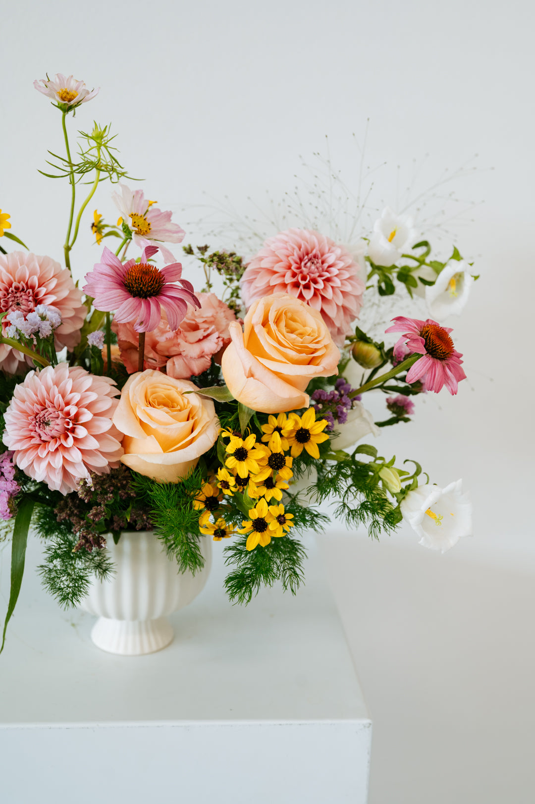 A pastel floral arrangement in a white ribbed ceramic vase, featuring peach roses, pink dahlias, yellow daisies, and green foliage, displayed on a white pedestal against a white background made by Flocal Co in Columbus, Ohio