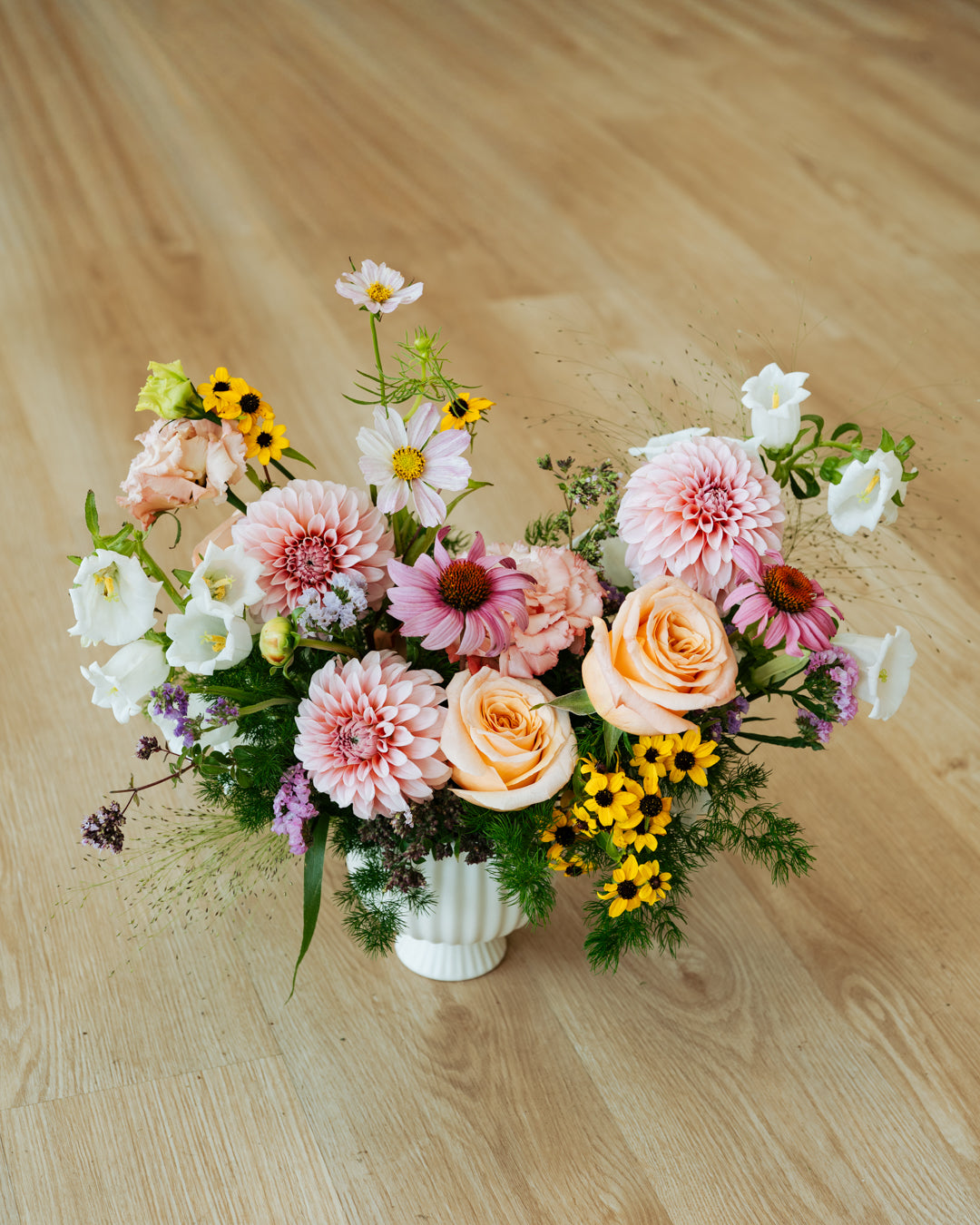 A vibrant, multicolored floral arrangement featuring pink and peach roses, dahlia, sunflowers, and white blossoms in a white ribbed ceramic pot, set on a light wood floor made by Flocal Co in Columbus, Ohio