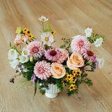 A vibrant, multicolored floral arrangement featuring pink and peach roses, dahlia, sunflowers, and white blossoms in a white ribbed ceramic pot, set on a light wood floor made by Flocal Co in Columbus, Ohio