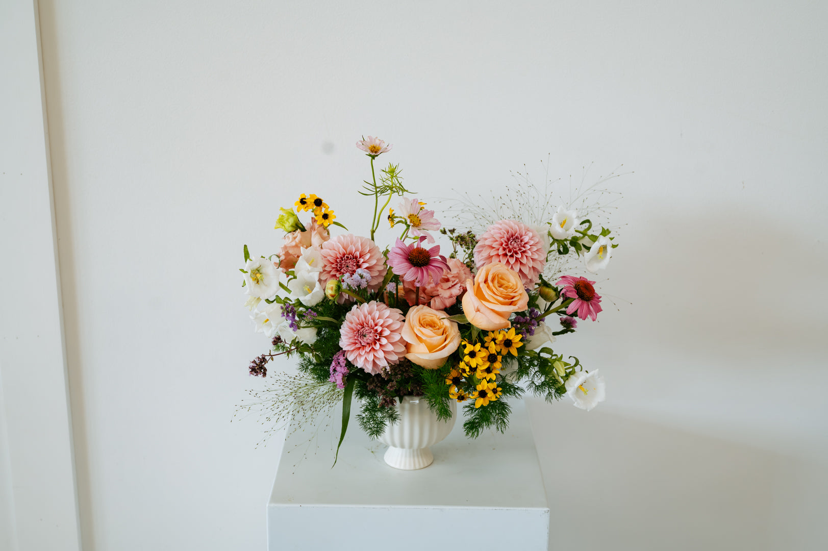 A vibrant floral arrangement featuring pink and peach roses, dahlia, yellow sunflowers, and green foliage in a white ribbed vase, displayed on a white pedestal against a plain white wall made by Flocal Co in Columbus, Ohio