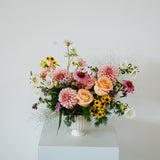 A vibrant floral arrangement featuring pink and peach roses, dahlia, yellow sunflowers, and green foliage in a white ribbed vase, displayed on a white pedestal against a plain white wall made by Flocal Co in Columbus, Ohio