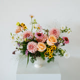 A pastel floral arrangement in a white ribbed vase, featuring pink and peach roses, dahlia-like blooms, yellow sunflowers, and green foliage, displayed on a white pedestal against a light gray background made by Flocal Co in Columbus Ohio