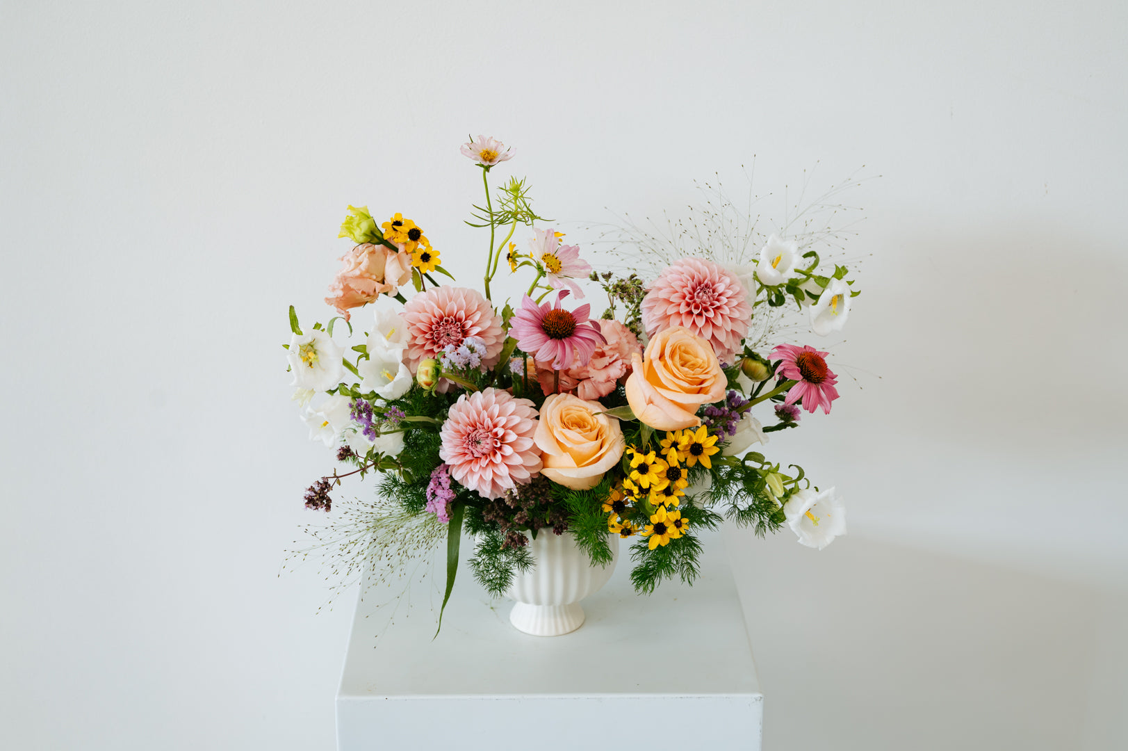 A pastel floral arrangement in a white ribbed vase, featuring pink and peach roses, dahlia-like blooms, yellow sunflowers, and green foliage, displayed on a white pedestal against a light gray background made by Flocal Co in Columbus Ohio