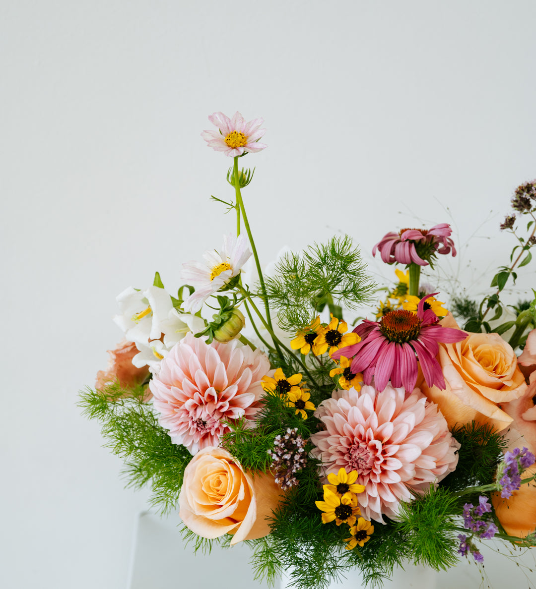 A vibrant, multicolored floral arrangement featuring pink, peach, yellow, and white flowers with green foliage, presented in a simple white cylindrical pot against a clean white background made by Flocal Co in Columbus, Ohio