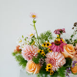 A vibrant, multicolored floral arrangement featuring pink, peach, yellow, and white flowers with green foliage, presented in a simple white cylindrical pot against a clean white background made by Flocal Co in Columbus, Ohio