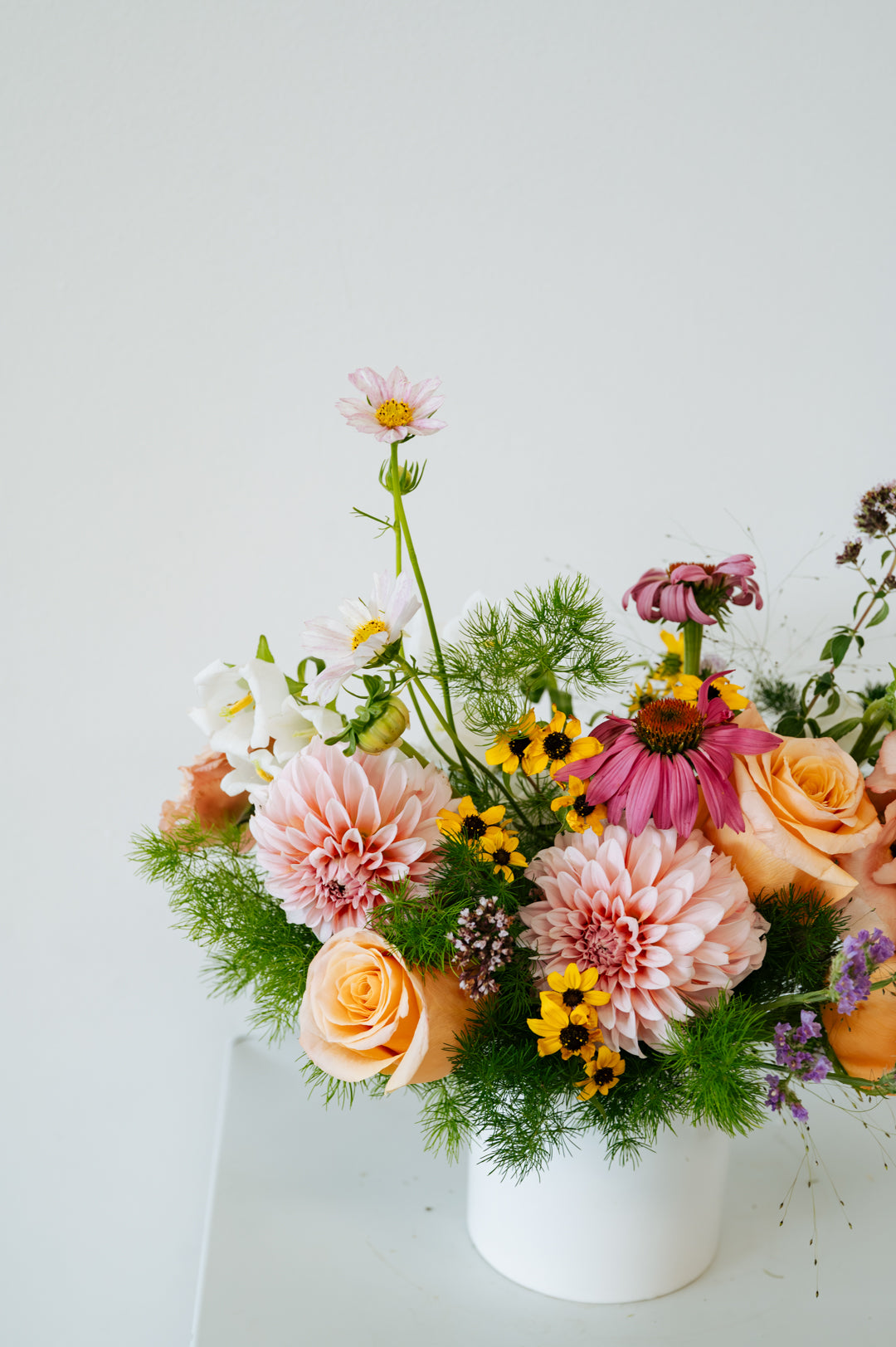 A vibrant, multicolored floral arrangement featuring pink, peach, yellow, and white flowers with green foliage, presented in a simple white cylindrical pot against a clean white background made by Flocal Co in Columbus, Ohio