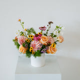 A colorful wildflower arrangement in a white ceramic pot, displayed on a white pedestal against a plain white wall made by Flocal Co in Columbus, Ohio 