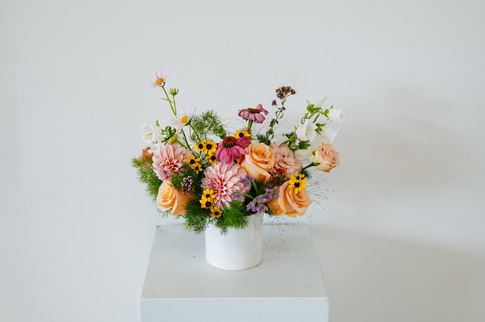 A colorful wildflower arrangement in a white ceramic pot, displayed on a white pedestal against a plain white wall made by Flocal Co in Columbus, Ohio 