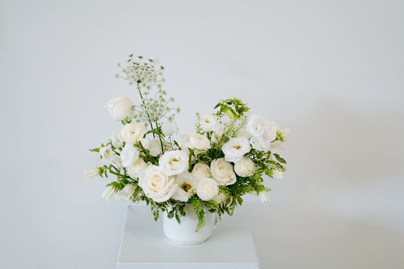 A white ceramic vase filled with a floral arrangement of white roses, white ranunculus, baby's breath, and green foliage, set against a plain white background.