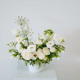 A white ceramic vase filled with a floral arrangement of white roses, white ranunculus, baby's breath, and green foliage, set against a plain white background.