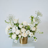 A white floral arrangement featuring roses, ranunculus, and baby's breath in a metallic gold cylindrical vase, displayed on a white pedestal against a light gray background available for local pick-up and delivery in Columbus, Ohio