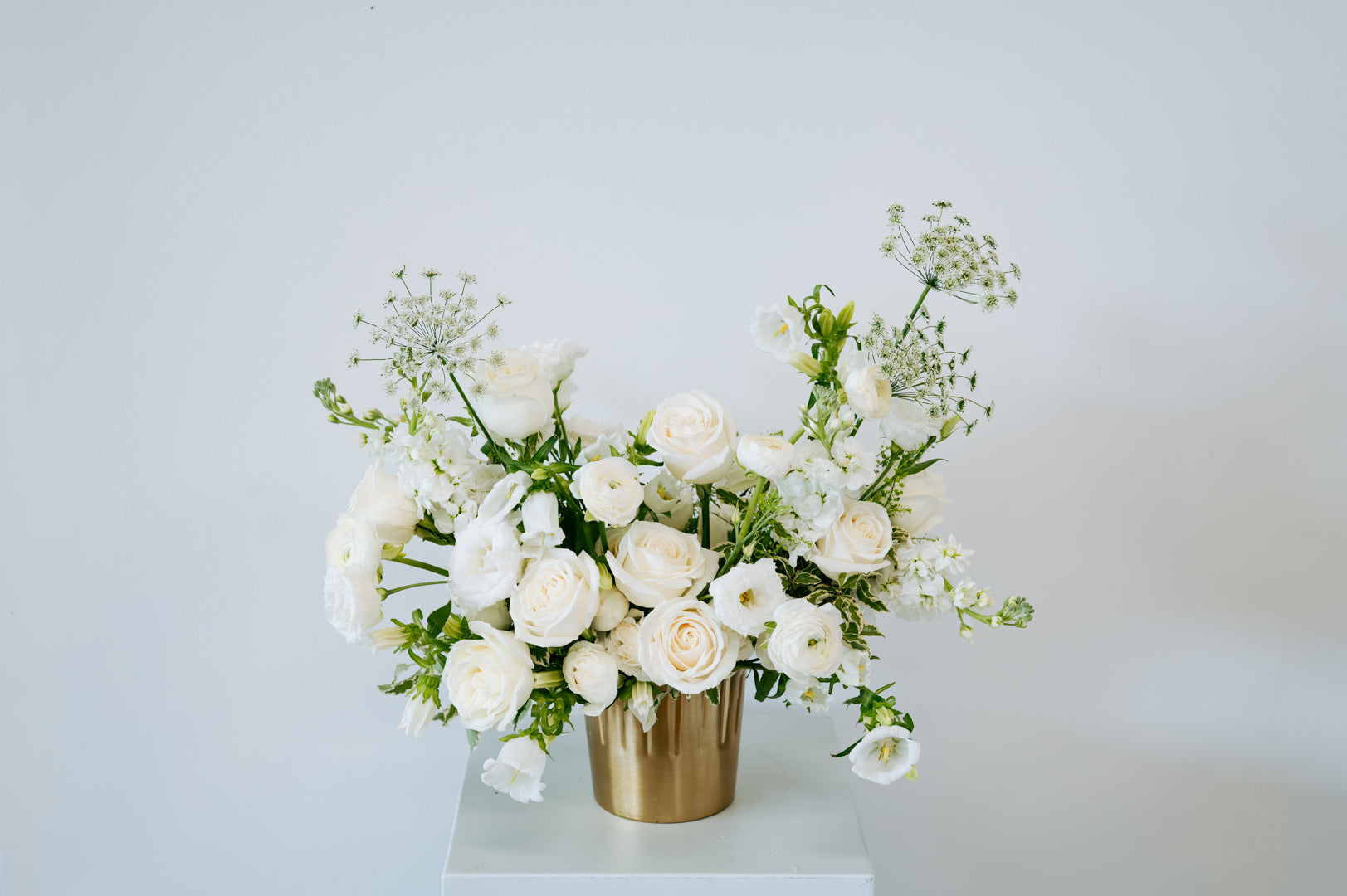 A white floral arrangement featuring roses, ranunculus, and baby's breath in a metallic gold cylindrical vase, displayed on a white pedestal against a light gray background available for local pick-up and delivery in Columbus, Ohio